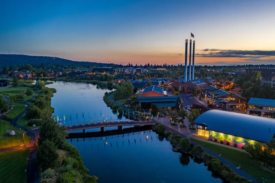Aerial view of Bend, Oregon with river in foreground and factory smokestacks with American flags on top against sunset sky.